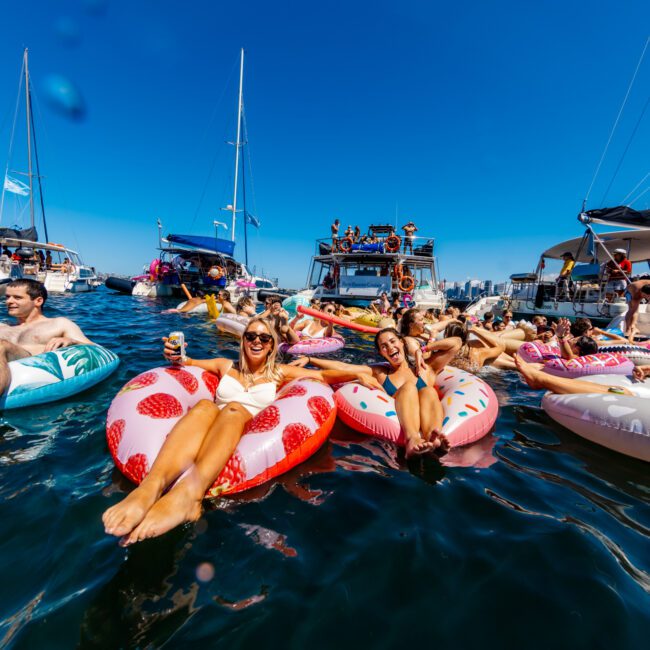 A group of people relax on colorful pool floats in the water near several boats. Two women in the foreground smile and enjoy drinks while sitting on strawberry and donut-shaped floats. The sunny day and clear blue sky add to the festive atmosphere at The Yacht Social Club Event Boat Charters.