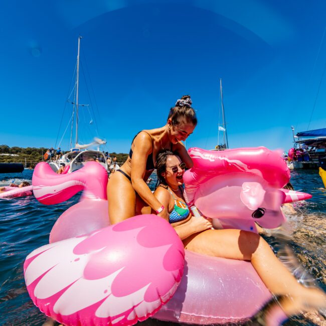 Two women enjoying a sunny day on the water, sitting on a large pink flamingo inflatable. They are in swimsuits, smiling and laughing, with boats and other people from The Yacht Social Club Sydney Boat Hire in the background. The sky is clear and blue, enhancing the vibrant and cheerful atmosphere.