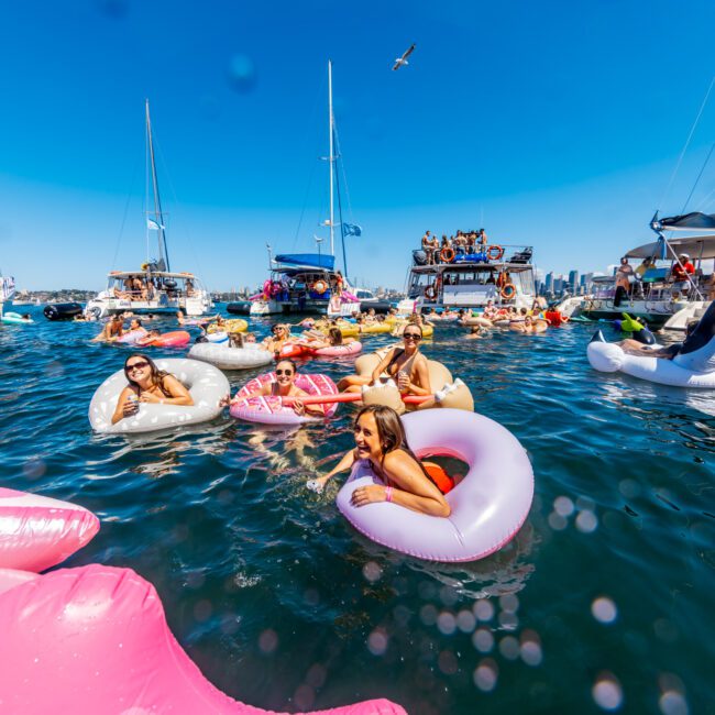 People float in colorful inflatable tubes in a vibrant, sunlit body of water, surrounded by several boats. A clear blue sky frames the scene with a seagull flying above. In the background, the silhouettes of buildings indicate an urban waterfront setting, perfect for a Luxury Yacht Rentals Sydney experience.