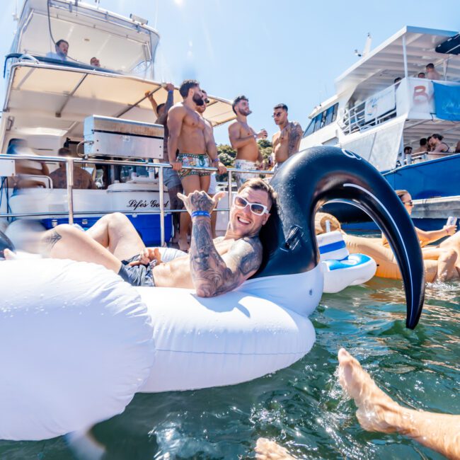 A man with tattoos and sunglasses lounges on an inflatable swan in the water, smiling and gesturing with his hand. In the background, people are socializing on a nearby boat from The Yacht Social Club Sydney Boat Hire. Several boats and flotation devices float around them on a sunny day.