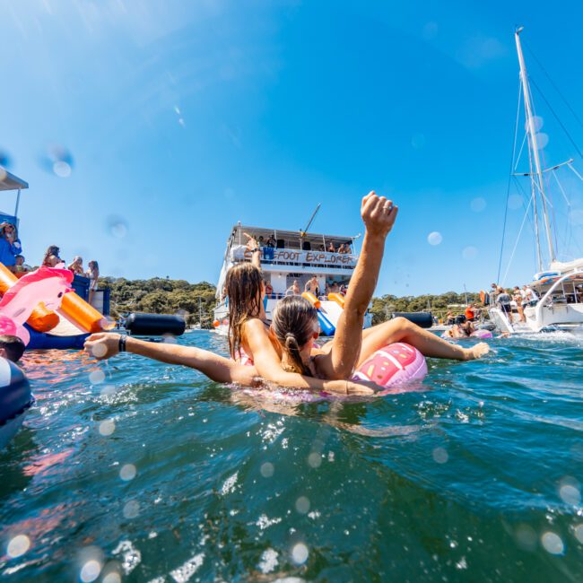 People enjoying a sunny day on the water, with some floating on inflatable rafts and others swimming near boats. The scene, part of The Yacht Social Club Sydney Boat Hire, includes a backdrop of a large boat and clear blue skies. The water is speckled with droplets, accentuating the lively atmosphere.