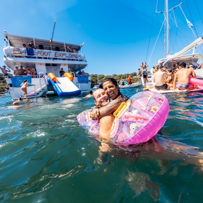 A joyful couple floats on a pink inflatable ring in the water, smiling at the camera. Behind them, people enjoy themselves on anchored boats from Boat Rental and Parties Sydney The Yacht Social Club. The sky is clear, and the water is vibrant under the bright sunlight.