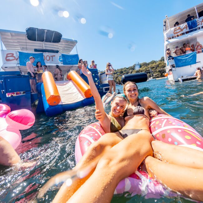 Two women in swimsuits are smiling and posing on a large inflatable float in the water. Several people are on nearby boats, with one boat having a large slide into the water. The sun is shining brightly, and the scene is lively with a festive atmosphere by The Yacht Social Club Event Boat Charters.