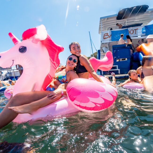 Two smiling women sit on a pink inflatable unicorn float at a sunny outdoor pool party. More people and various colorful floats are visible in the background alongside The Yacht Social Club Event Boat Charters. The scene is vibrant and festive with a large boat and bright blue sky completing the atmosphere.