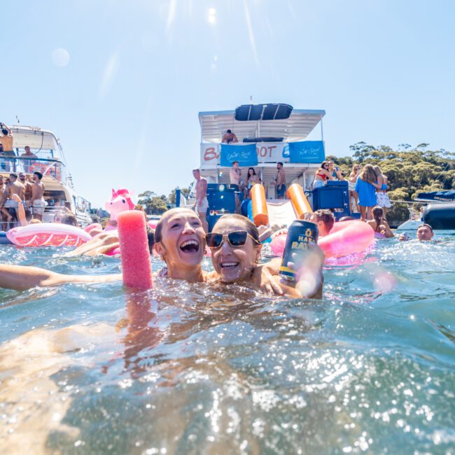 Two smiling individuals are enjoying time in the water near a boat, holding drinks and floating on colorful inflatables. They are surrounded by several other people on boats in the background, under a clear blue sky. The atmosphere at Sydney Harbour Boat Hire The Yacht Social Club is lively and joyful.