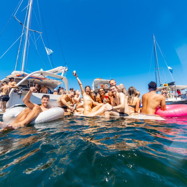 A group of people in swimsuits enjoying a sunny day on the water near boats. They are sitting on inflatable floats and holding drinks, with others swimming around. Bright blue sky and sailboats are visible in the background, a perfect scene typical of The Yacht Social Club events by Sydney Harbour Boat Hire.