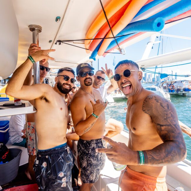 A group of men enjoying a sunny day on a boat. They are shirtless, smiling, and striking playful poses. The boat, provided by Luxury Yacht Rentals Sydney's Yacht Social Club, is equipped with colorful pool noodles and docked near other vessels. The sky is clear and the water calm, perfect for boating.