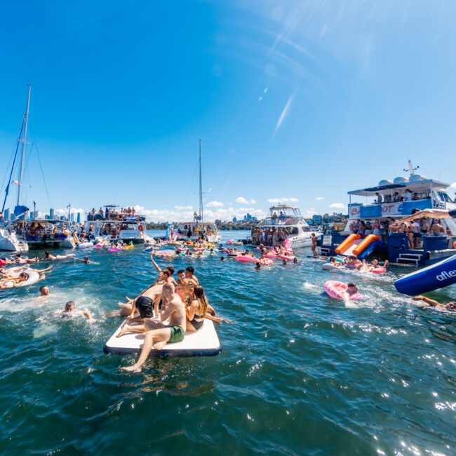 People enjoy a sunny day on the water with boats, yachts, and inflatables. Some are swimming, while others relax on floating devices. Boats from The Yacht Social Club Event Boat Charters are anchored nearby, and a city skyline can be seen in the background against a clear blue sky.