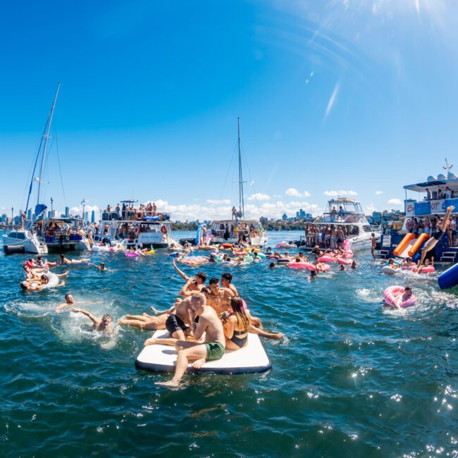 A lively scene of people enjoying a sunny day on the water. Numerous boats are anchored close together with partygoers from The Yacht Social Club swimming, sunbathing, and socializing on floaties and inflatables. The city skyline is visible in the distance under a clear, blue sky.