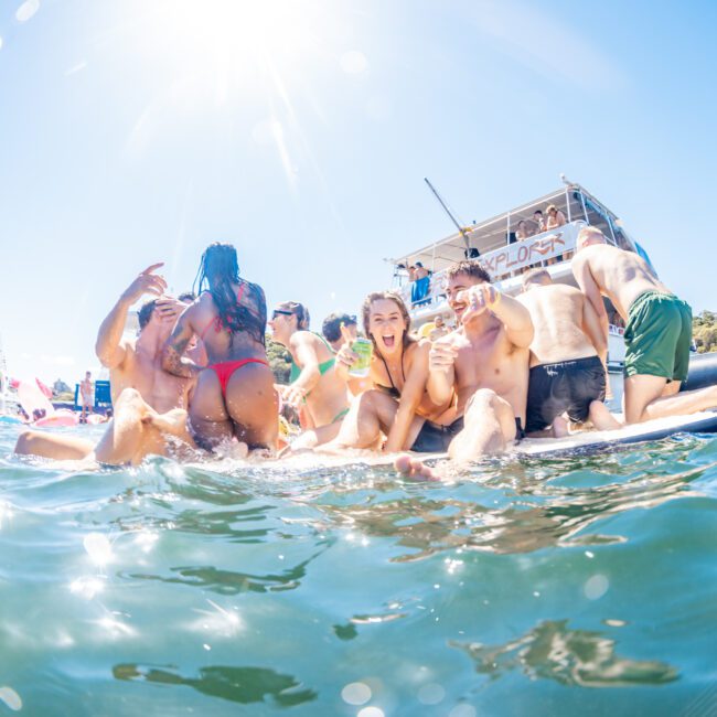 A group of people in swimwear enjoy a sunny day on the water, some embracing and laughing on an inflatable raft. A boat with the word 'EXPLORER' is seen in the background, adding to the festive and lively setting. It's a perfect day for The Yacht Social Club Sydney Boat Hire experience.