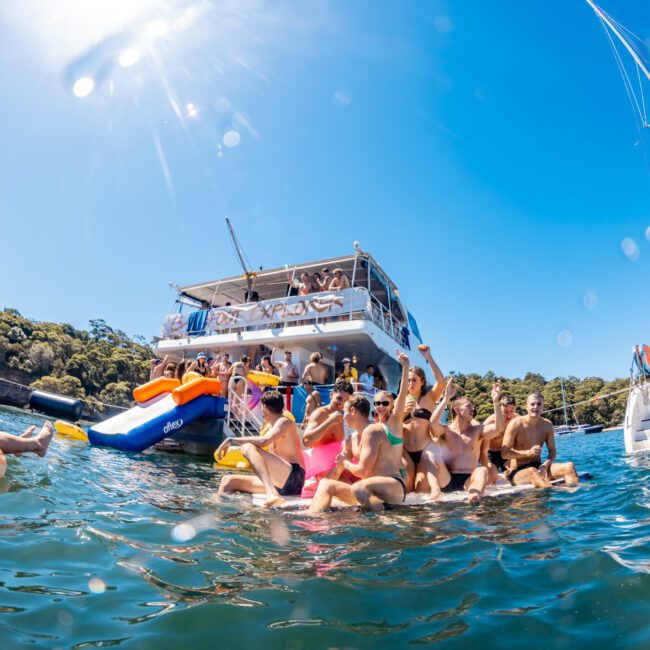 A group of people enjoying themselves in the water near a boat on a sunny day, with The Yacht Social Club Sydney Boat Hire. The boat has two levels, and more people are seen on the upper deck. Another boat is visible in the background, and the water is calm with light reflections. Trees are in the distance.