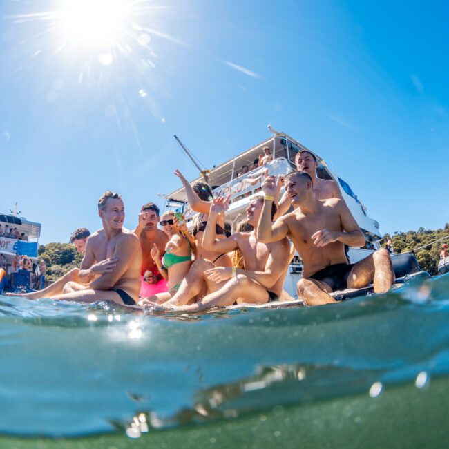 A group of people in swimwear are enjoying a sunny day in the water near a boat from The Yacht Social Club. They are smiling, laughing, and splashing water. The sun is shining brightly in the clear blue sky, and the background features another boat and some greenery.