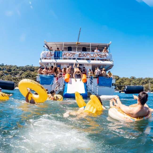 A lively scene of The Yacht Social Club Event with numerous people enjoying the water. Partygoers on colorful inflatables float near two large boats, with more revelers on the decks. The sun is shining, the sky is clear, and there are trees visible in the background.