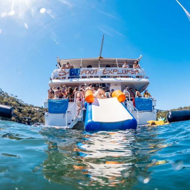 A group of people enjoy a sunny day on a large boat named "Rocky Foot Explorer." Some are standing on the deck, while others slide down a white and blue inflatable slide into the water. The boat, part of The Yacht Social Club Event Boat Charters, floats near a wooded shoreline under a clear, blue sky.