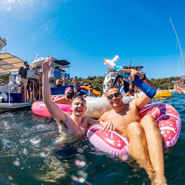 Two men are smiling and enjoying themselves on inflatable floats in the water, surrounded by boats and other people in the background. One man is holding a bottle and the other is raising his hand in excitement. It’s a sunny day with a clear blue sky, perfect for The Yacht Social Club Sydney Boat Hire adventure.