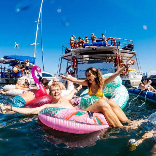 A lively group of people enjoying a sunny day on the water, floating on colorful inflatable pool toys near several anchored boats. Some are laughing and raising their arms, while others relax and socialize. The Yacht Social Club Event creates a festive atmosphere with the bright blue sky and clear water.