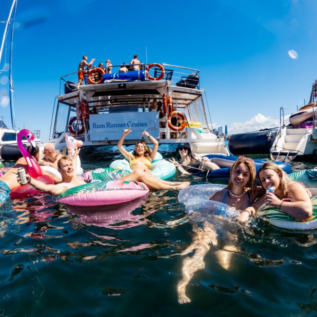 A group of people is enjoying a sunny day on the water, lounging on colorful inflatable pool floats shaped like a flamingo and a donut near two boats. The Yacht Social Club Sydney Boat Hire creates a lively and cheerful atmosphere under the clear blue sky with calm water.