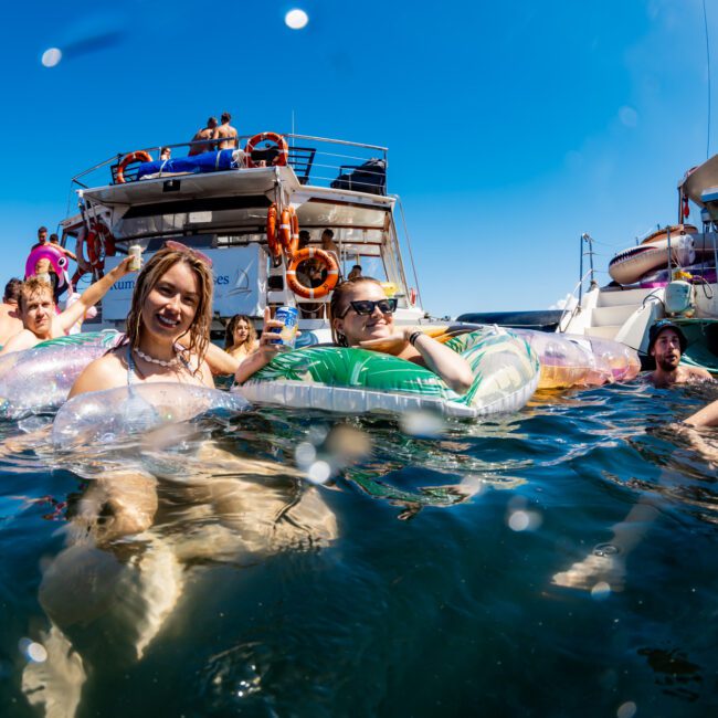 A group of people enjoying a sunny day on Sydney Harbour Boat Hire The Yacht Social Club, floating on colorful inflatables. They are smiling, wearing swimsuits, and relaxing in the water. The sky is clear and bright blue, creating a vibrant and joyful atmosphere.