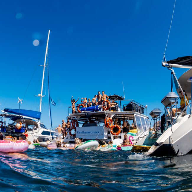 A lively scene of people enjoying a day on the water, with several boats gathered close together from The Yacht Social Club. Individuals are swimming, floating on inflatables, and lounging on boats under a clear blue sky. There is an atmosphere of fun and leisure.