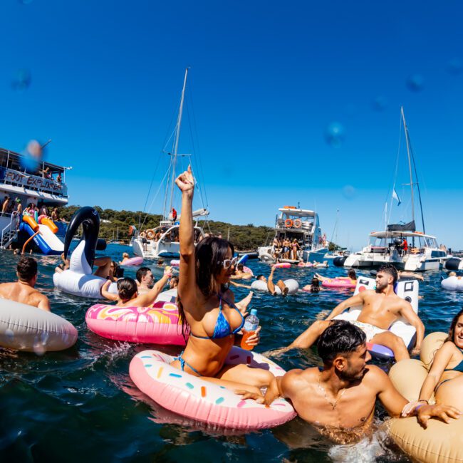 A lively scene of people enjoying a sunny day on the water, some floating on inflatable tubes and others swimming. Boats and yachts hired from Sydney Harbour Boat Hire The Yacht Social Club are anchored in the background, and the blue sky is clear, indicating a warm and cheerful atmosphere.