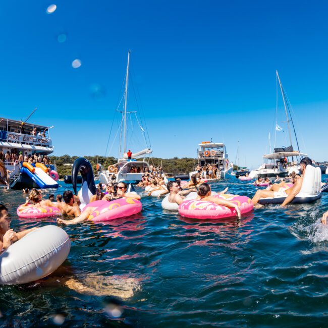 People are enjoying a sunny day on the water surrounded by several boats. They are floating on inflatable pool toys shaped like donuts and other fun shapes. Some partygoers with Sydney Harbour Boat Hire The Yacht Social Club are dancing and socializing on the boats in the background under a clear blue sky.