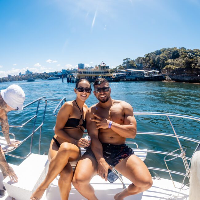 A group of people enjoys a sunny day on a boat. A man and woman, both in swimwear, sit at the front smiling and posing for the camera. Behind them, someone with tattoos bends over. The water and a shoreline with trees and buildings are visible in the background, showcasing The Yacht Social Club Sydney Boat Hire.