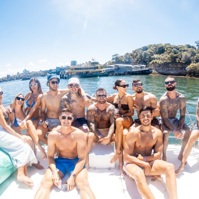 A group of 12 people, wearing swimsuits, are sitting together on a luxury yacht under a sunny, clear blue sky. They are smiling and posing for the camera. The water and shoreline, featuring trees and buildings, are visible in the background—an ideal setting for Boat Parties Sydney The Yacht Social Club.