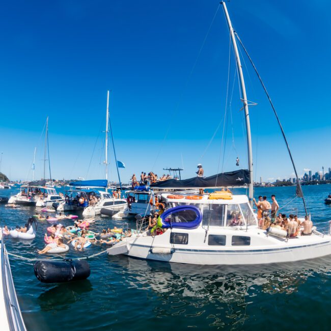 A vibrant scene on the water features several boats gathered together in a party atmosphere. People are sunbathing, swimming, and lounging on inflatables. The skyline of Sydney is visible in the background under a clear blue sky, courtesy of Boat Rental and Parties Sydney - The Yacht Social Club.