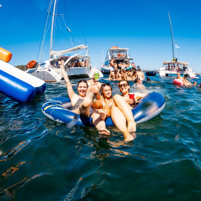 Three people relax on inflatable rafts in the water, smiling and enjoying beverages, surrounded by boats and other people in the background. The scene is bright and lively under a clear blue sky, embodying The Yacht Social Club Sydney Boat Hire's vibrant party atmosphere.