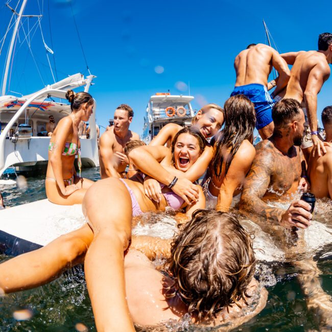 A lively group in swimwear enjoy a sunny day on a luxury yacht, laughing and swimming. Some are onboard, others in the water, while a few jump in, creating an energetic scene. A clear blue sky and other boats are visible in the background—courtesy of The Yacht Social Club Sydney Boat Hire.