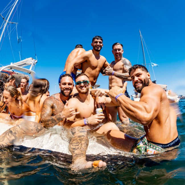 A group of young people are enjoying a sunny day on The Yacht Social Club Event Boat Charters. Some are sitting on the boat, while others are in the water. They are smiling, holding drinks, and appear to be having a fun time. The background shows clear blue skies and other boats nearby.