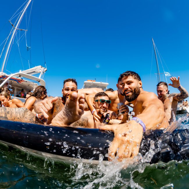 A group of people in swimwear are having fun on an inflatable raft in the water with boats visible in the background. They are smiling, splashing water, and posing for the camera on a sunny day under a clear blue sky. It's a perfect scene for The Yacht Social Club Sydney Boat Hire experience.