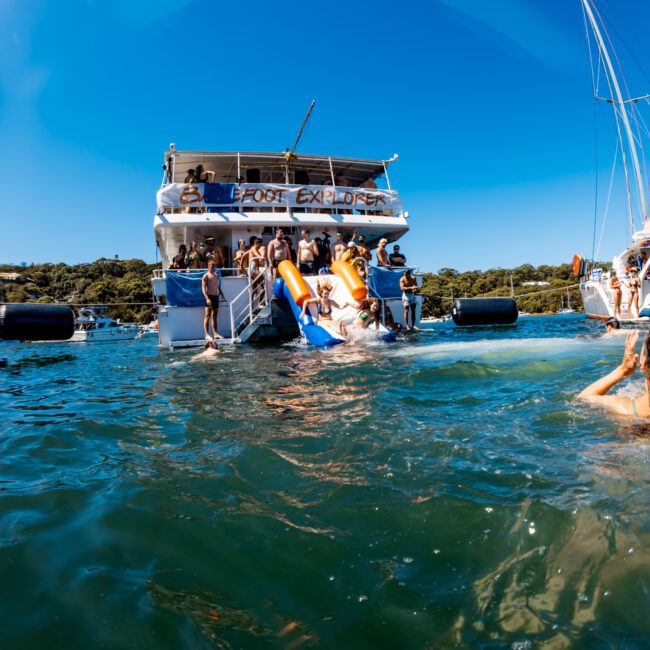 A lively scene of people enjoying water activities near a boat on a sunny day. Several individuals slide down a water slide from the boat into the sea while others swim and play around. The colorful luxury yacht, part of The Yacht Social Club Event Boat Charters, has trees and clear skies in the background.