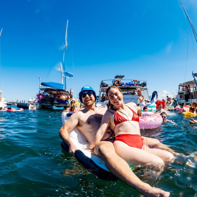 A group of people enjoy a sunny day on the water. In the foreground, a smiling man in sunglasses and a woman in a red bikini relax on a float. Boats from The Yacht Social Club Event Boat Charters and more people on floats can be seen in the background. The sky is clear and blue.