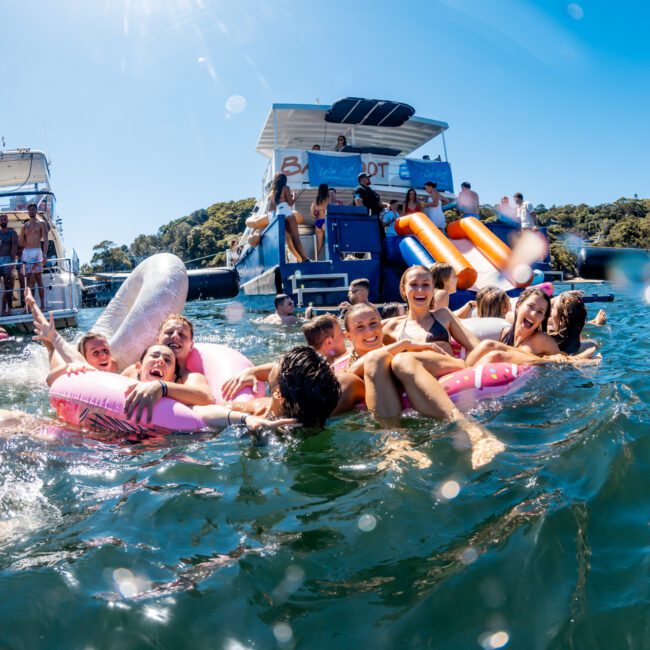 A group of people are enjoying a sunny day in the water, floating on inflatable rafts and ring tubes. Boats from The Yacht Social Club Sydney Boat Hire are anchored nearby, with more people on board. Some boats have slides, and everyone seems to be having fun against a backdrop of clear blue skies and greenery.