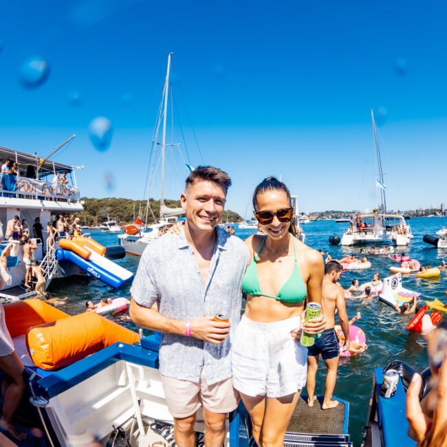 A man and woman smile for a photo on a boat at a lively party hosted by The Yacht Social Club Sydney Boat Hire. They are surrounded by people in swimsuits, inflatables, and boats on a sunny day. The woman wears a green bikini top and white shorts, while the man wears a light blue shirt and shorts, both holding drinks.