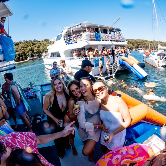 A lively boat party with people enjoying themselves on a sunny day. Three women in the foreground smile and pose with drinks. In the background, others swim, float on inflatables, and socialize on two large boats anchored close to each other at The Yacht Social Club Event Boat Charters.