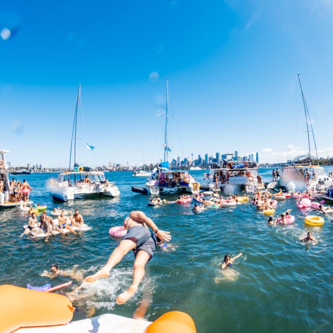 A vibrant floating party from The Yacht Social Club is in full swing on a sunny day, with numerous people swimming and floating on inflatables between sailboats and yachts. In the foreground, a person is mid-dive into the water. Sydney's city skyline and clear blue sky are visible in the background.