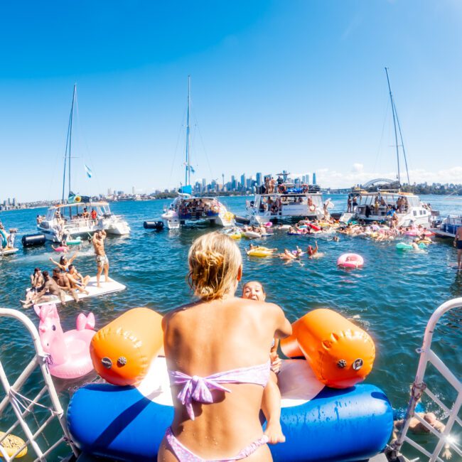 A vibrant aquatic scene shows people enjoying a sunny day on a lake or coastal area. Numerous boats, including luxury yacht rentals from The Yacht Social Club Sydney, are docked close together, with people swimming, floating, and lounging on inflatable toys. The clear blue sky and distant city skyline create a lively atmosphere.