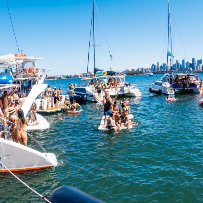 A lively scene of people enjoying a sunny day on boats and yachts in a large body of water. Many are swimming, lounging, and socializing as part of The Yacht Social Club Sydney Boat Hire. The city skyline is visible in the background.