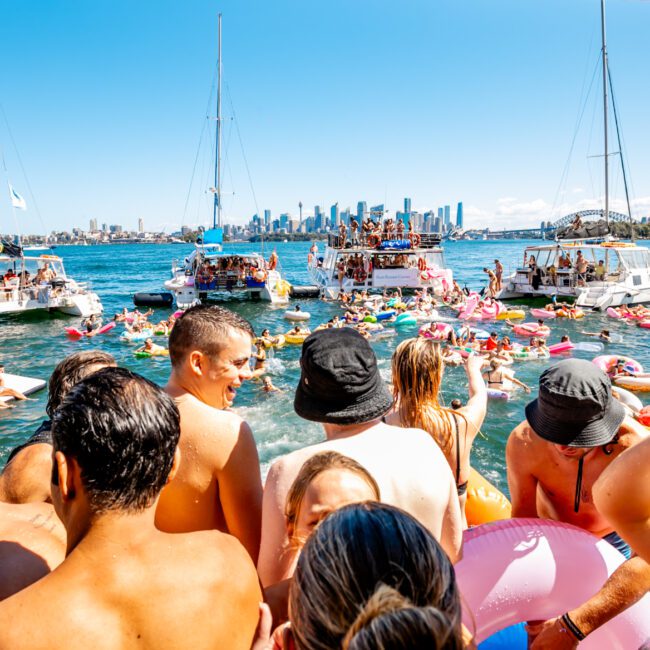 A lively boat party on a sunny day in Sydney Harbour, hosted by The Yacht Social Club Event Boat Charters, with the city skyline in the background. People swim and lounge on various inflatables in the water, as several boats are anchored close together creating a festive atmosphere.