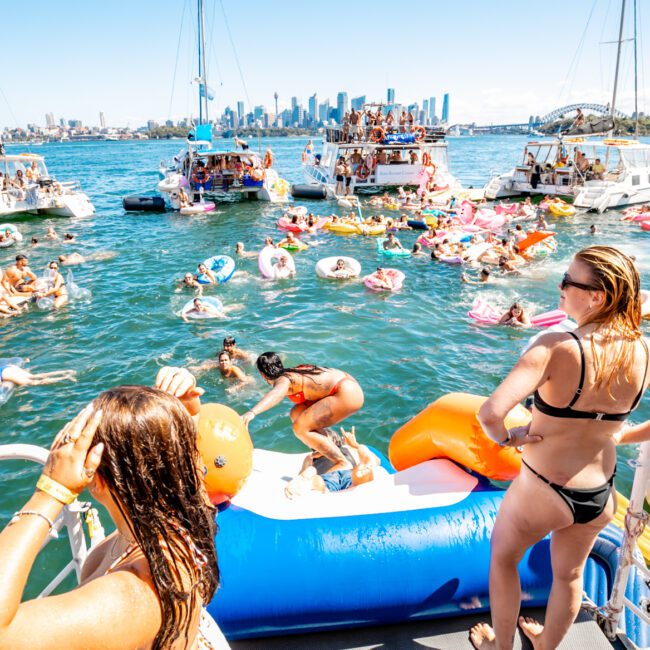 People enjoy a sunny day on the water near a city skyline, with groups swimming, lounging on boats, and using inflatable floats. Several yachts from The Yacht Social Club Sydney Boat Hire form a semi-circle, creating a lively and relaxed atmosphere.