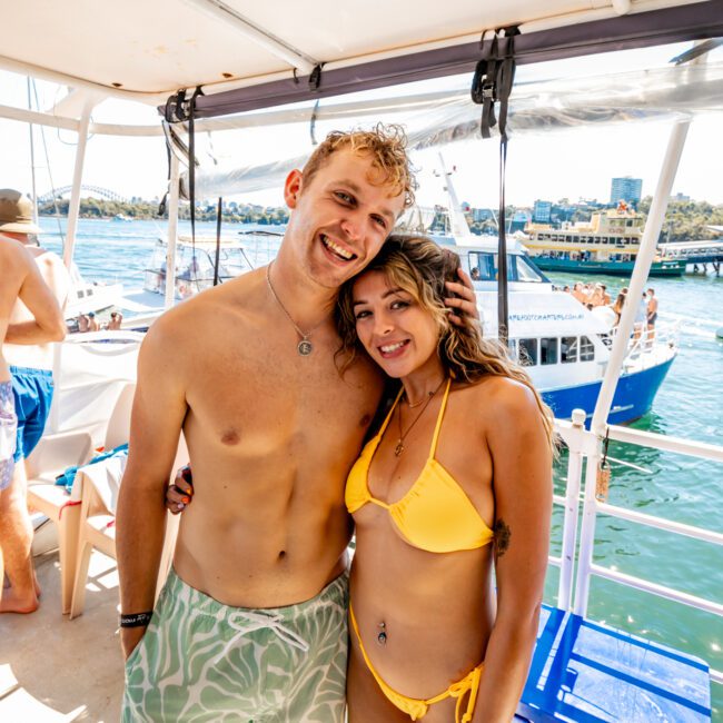 A shirtless man and a woman in a yellow bikini stand closely together on a boat, both smiling at the camera. In the background, other people, a blue boat, and the city skyline with a bridge over water on a sunny day can be seen. Enjoy Luxury Yacht Rentals Sydney with The Yacht Social Club for an unforgettable experience.