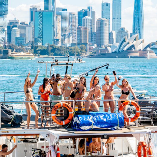 A group of people in swimwear dance and cheer on the upper deck of a boat labeled "Rum Runner Cruises," with a backdrop of a city skyline featuring modern skyscrapers, the Sydney Opera House, and the waters of Sydney Harbour sparkling under a clear blue sky. It's all part of The Yacht Social Club's vibrant boat parties in Sydney.