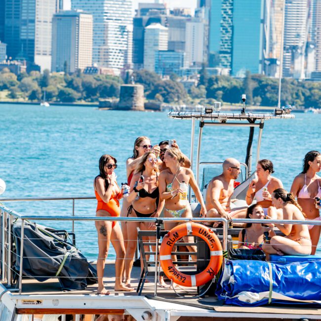 A group of people enjoying a sunny day on a boat with a city skyline in the background. Some are standing and taking pictures, while others are sitting and chatting. Everyone appears relaxed and dressed in swimwear, indicating a casual and festive atmosphere aboard The Yacht Social Club's luxury yacht rental in Sydney Harbour.