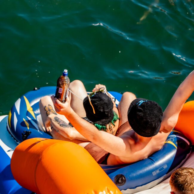 Two individuals are lounging on an inflatable raft in the water, one holding a drink bottle. They are wearing hats and swimwear. Other people are swimming in the background. The scene suggests a sunny day at a lake or seaside, reminiscent of a day with The Yacht Social Club Sydney Boat Hire.