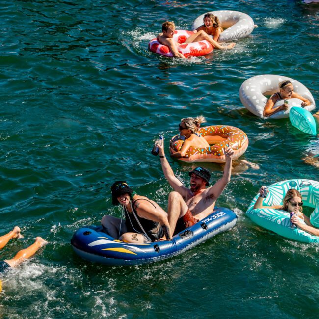 A group of people floating on a river in colorful inflatable tubes and rafts. Some are laughing and holding drinks, while others are relaxing and enjoying the sunny day. The clear, vibrant blue water adds to the lively, festive scene, reminiscent of a Sydney Harbour Boat Hire event by The Yacht Social Club.