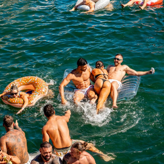 A group of people are enjoying a sunny day on the water, some floating on inflatables and others swimming. Some inflatables are donut-shaped, and there's a floating mat as well. Everyone appears to be having fun, interacting playfully and relaxing in the water—almost like a luxury yacht rental in Sydney.