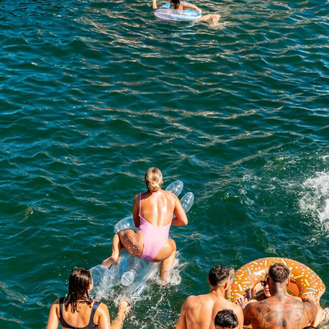 A group of people enjoy a sunny day by the water. In the foreground, a woman rides an inflatable floatie shaped like a donut while others look on with excitement. In the background, someone paddles on an inflatable ring. Everyone is wearing swimwear, embracing the fun vibe of The Yacht Social Club Sydney Boat Hire.