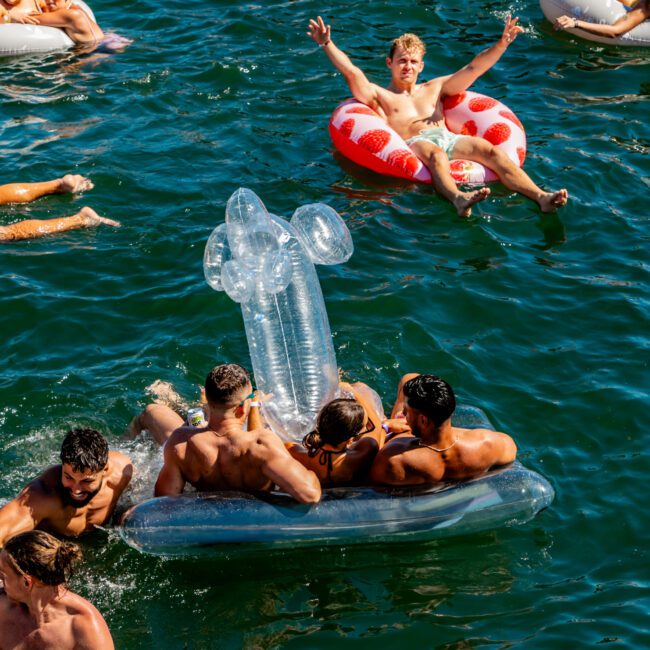 A group of people enjoying a sunny day on the water. Some are relaxing on inflatable floats, including a clear float with four people and a red and white float with a person raising their arms. The Yacht Social Club Event Boat Charters offer vibrant blue waters where everyone seems to be having fun.